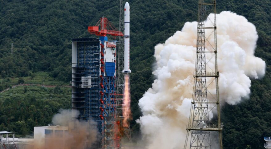 A 41-meter-long white Long March 2D rocket rises from its launch tower at hill-surrounded Xichang launch center. Mach diamonds form in the exhaust while plumes of white smoke billow away from the path of the rocket.