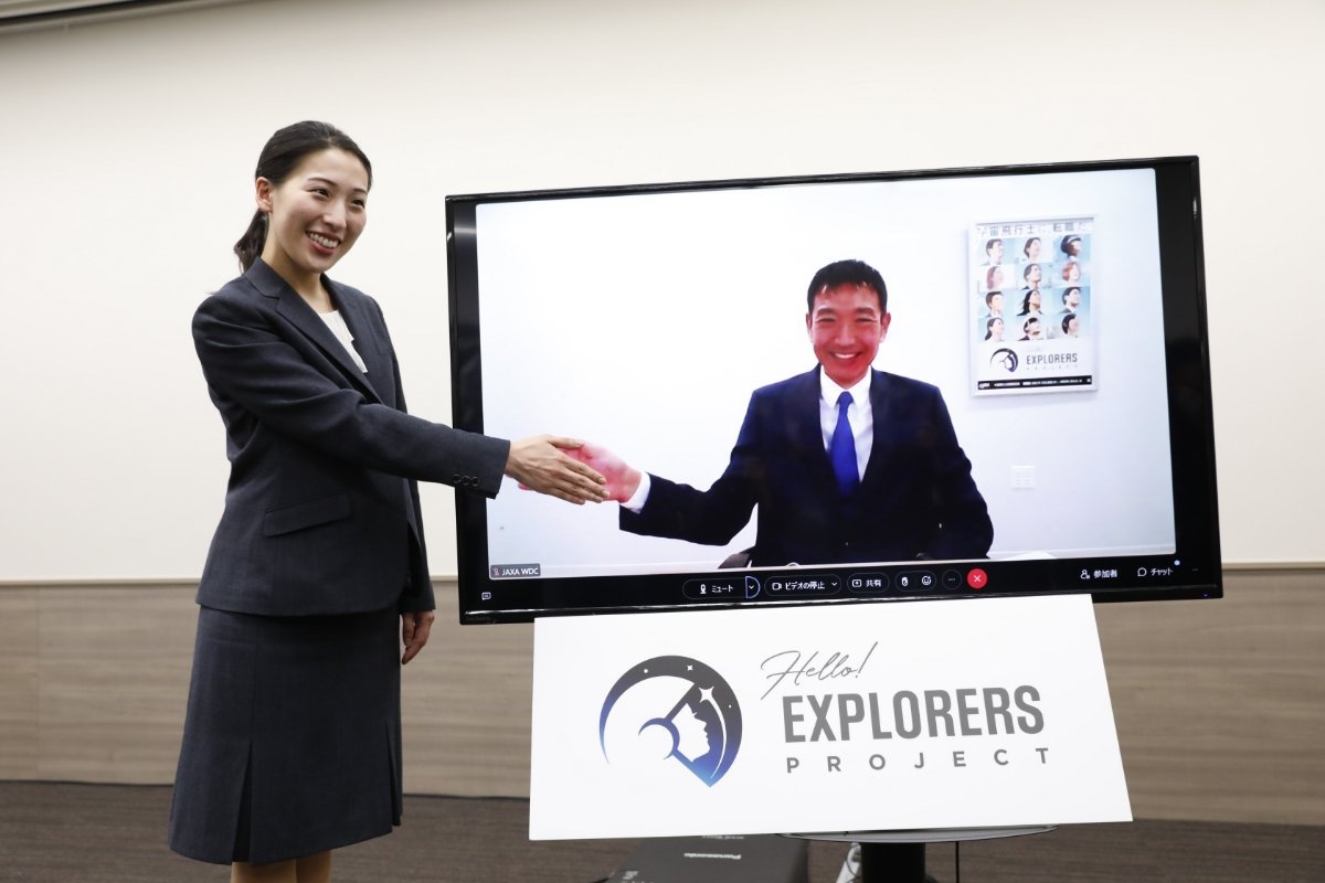 A Japanese woman in a gray business suit smiles as she pretends to shake hands with a Japanese man on a television screen.