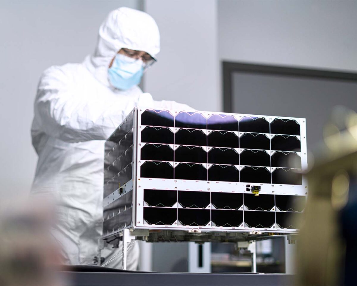 a technician in a white "bunny suit" works on a rectangular nanosatellite the size of a microwave oven.