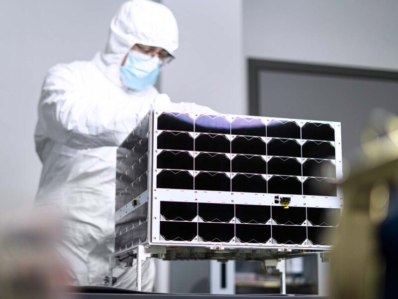 a technician in a white "bunny suit" works on a rectangular nanosatellite the size of a microwave oven.