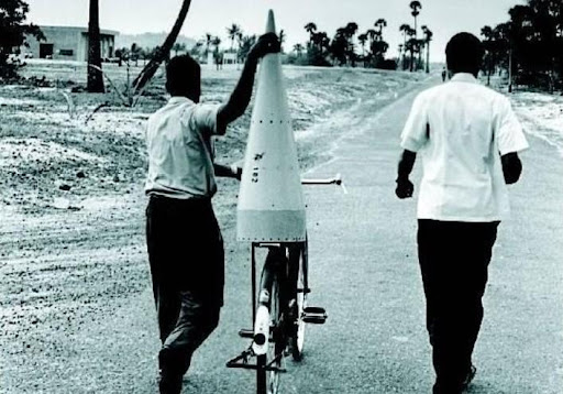 black and white photo of two men transporting a rocket cone supported on a bicycle.